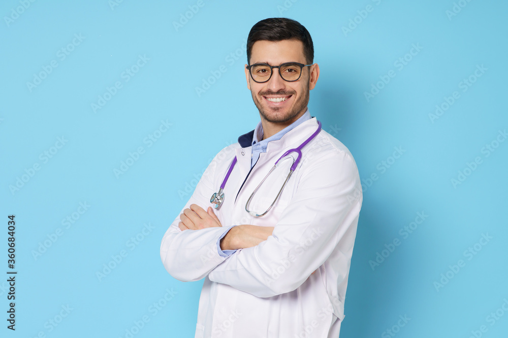 Young medical guy dressed in white doctor robe with stethoscope round his neck, holding arms crossed, smiling happily, isolated on blue background Young medical guy dressed in white doctor robe with stethoscope round his neck, holding arms crossed, smiling happily, isolated on blue background