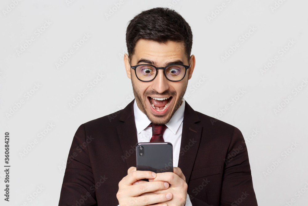 Young business man looking at smart phone with surprise expression, shouting WOW, isolated on gray background Young business man looking at smart phone with surprise expression, shouting WOW, isolated on gray background