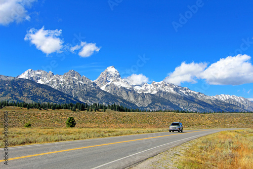 Beautiful scenic drive in Grand Teton National park - isolated car driving on the empty road / highway surrounded by high snowy rocky mountain peaks and fields. Wyoming, USA.