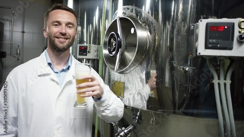 A happy dark-haired bearded laboratory worker in a white overall is smiling and holding the glass fith the final product of a brewery photo