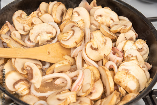 Cooking the squid and mushroom sauce. Sliced squid rings and champignons are stewed in a pan and mixed with a wooden spoon.