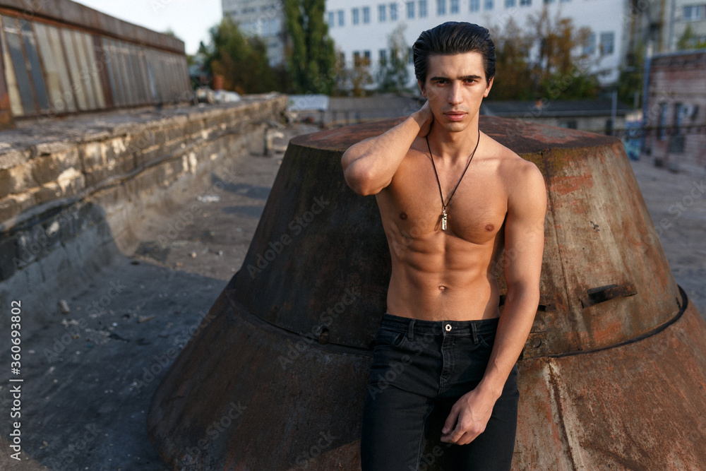 Close up portrait of a athletic young man with naked torso posing on the roof at the sunset background.