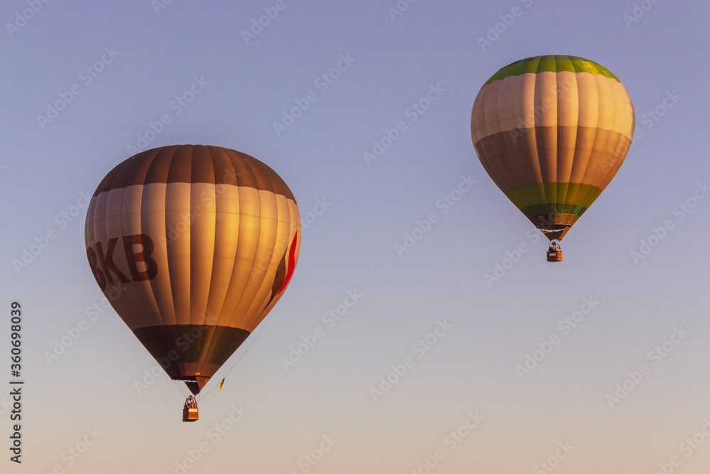 Naklejka premium Kyiv, Ukraine - 06/27/2020: Hot air balloons in clear blue sky. Colorful balloons with basket lift up. Summer adventure and leisure. Aviation sport concept. Two balloons in morning light.