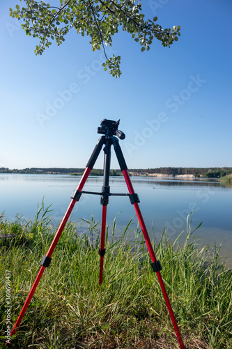 Camera red tripod standing in green grass near lake water natural landscape on clear sunny bright day