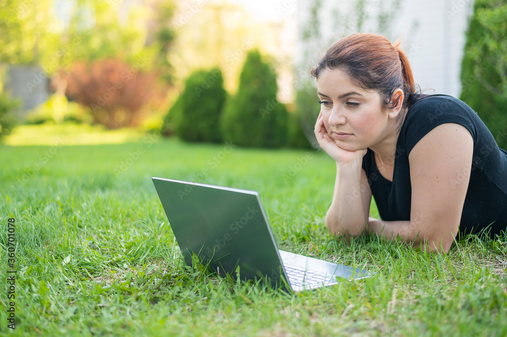 A woman lies on her stomach on the lawn at the cottage and uses on a laptop. A focused female freelancer works remotely outdoors. The girl is studying on the computer. A woman lies on her stomach on the lawn at the cottage and uses on a laptop. A focused female freelancer works remotely outdoors. The girl is studying on the computer.
