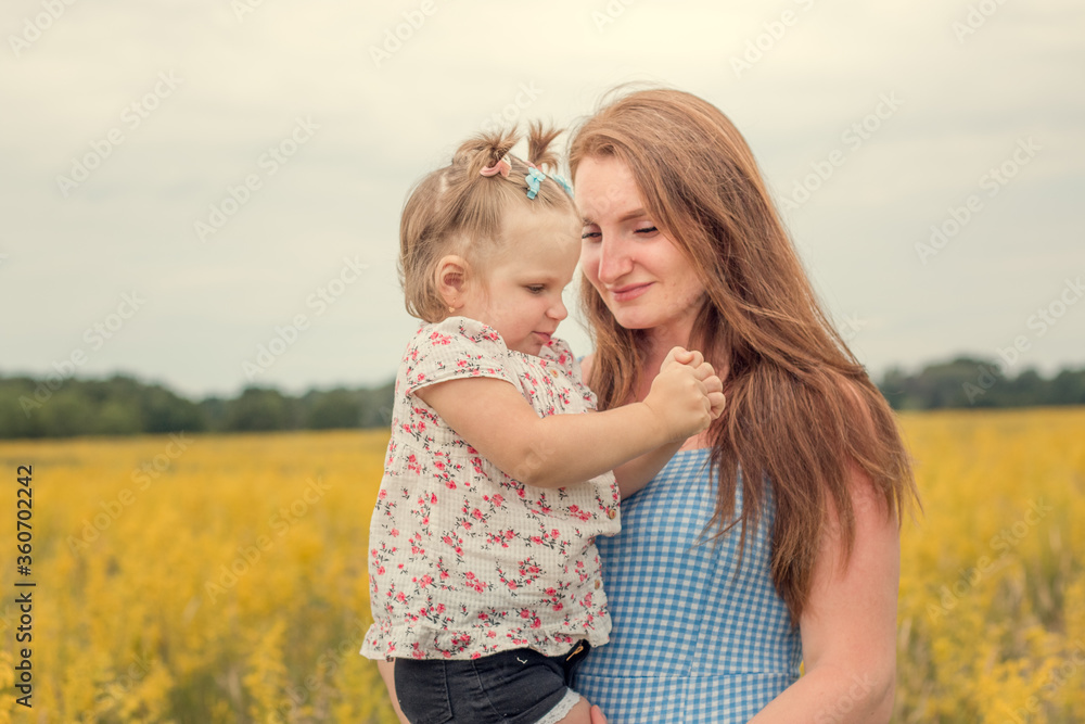 A little beautiful happy girl in her mother's arms. In everyday clothes. Smiling in a yellow field. With ponytails on their heads. Horizontal photo. Cloudy weather. Summer. A little beautiful happy girl in her mother's arms. In everyday clothes. Smiling in a yellow field. With ponytails on their heads. Horizontal photo. Cloudy weather. Summer.