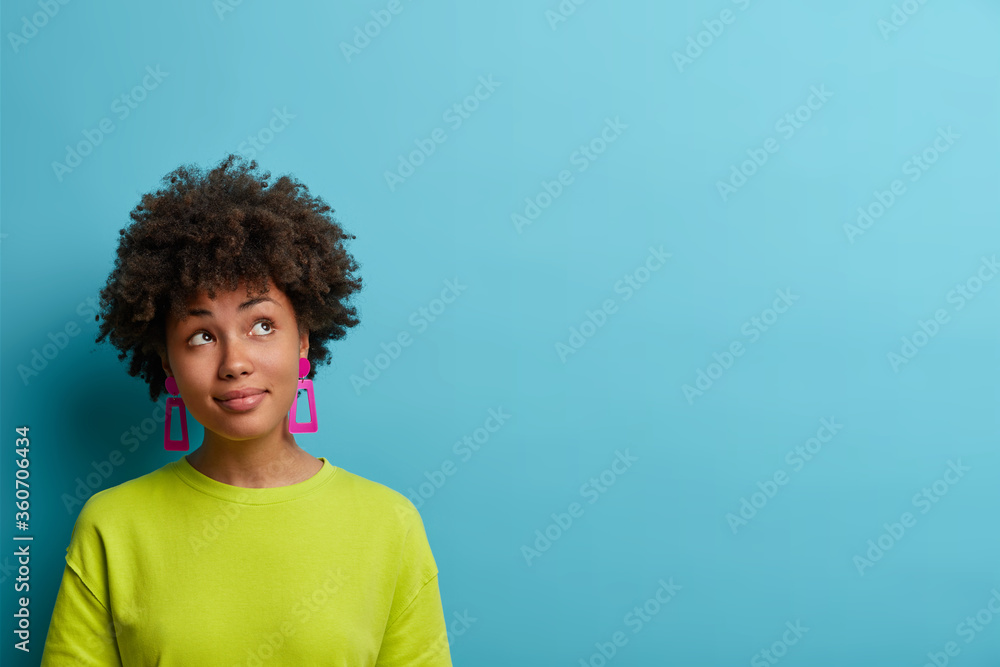 Photo of serious dreamy woman with bushy curly hair, concentrated above and ponders on nice plan, has satisfied expression, wears bright summer clothes, models against blue background, copy space Photo of serious dreamy woman with bushy curly hair, concentrated above and ponders on nice plan, has satisfied expression, wears bright summer clothes, models against blue background, copy space