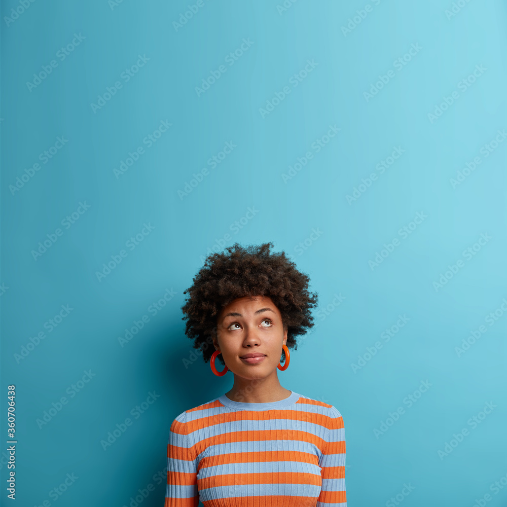Vertical shot of cute dark skinned woman with Afro hairstyle, looks above with pondering expression, notices something interesting, blank space on blue background. Face expressions and emotions - obrazy, fototapety, plakaty 