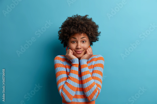 Portrait of beautiful curly woman with natural beauty, keeps both hands under chin, listens interlocutor with interest, has gentle smile, dressed in striped jumper, isolated over blue background