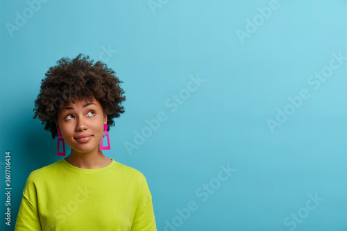 Photo of serious dreamy woman with bushy curly hair, concentrated above and ponders on nice plan, has satisfied expression, wears bright summer clothes, models against blue background, copy space