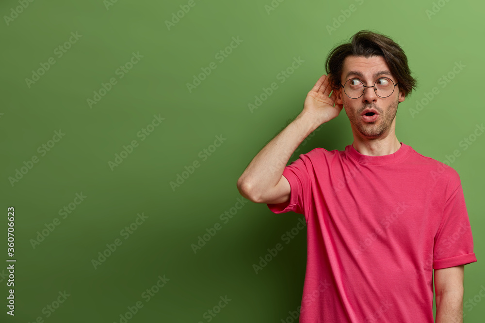 Surprised wondered guy tries to overhear some news, keeps hand near ear, puts nose in business of other people, tries to hear some information and wants to spread rumors, stands over green wall Surprised wondered guy tries to overhear some news, keeps hand near ear, puts nose in business of other people, tries to hear some information and wants to spread rumors, stands over green wall