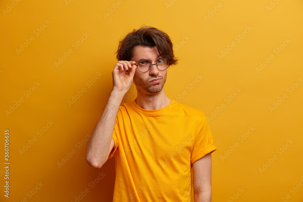 Portrait of adult man keeps hand on rim of round spectacles, looks scrupulously and attentive, explores something, wears yellow t shirt, poses indoor, has ambitious plans for distant future. Portrait of adult man keeps hand on rim of round spectacles, looks scrupulously and attentive, explores something, wears yellow t shirt, poses indoor, has ambitious plans for distant future.