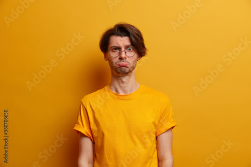 Photo of distressed unhappy European man purses lips, has offended sad face expression, resentful face, bad mood, looks through round spectacles, wears yellow t shirt, poses indoor. Human emotions © wayhome.studio 