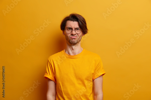 Half length shot of displeased man purses lower lip, looks offended and unhappy, finds out about his failure, has pessimistic attitude to everything, wears glasses and t shirt, poses over yellow wall © wayhome.studio 