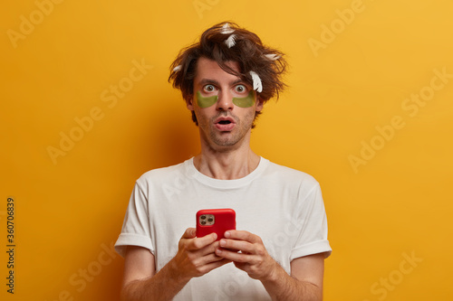 Shocked adult man stares at camera with fearful expression, checks email box, surfs social networks after awakening while undergoes facial beauty treatment, applies patches for reducing wrinkles