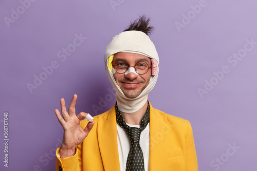 Funny glad man with shiner and broken nose, wears bandage on head, makes okay gesture, assures that everything is okay, smiles pleasantly, forgets about troubles and all his injuries has positive look photo