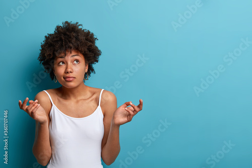 Clueless perplexed dark skinned woman with curly hair shrugs shoulders and looks questioned upwards, raises hands in hesitation, dressed casually, poses against blue studio background, copy space