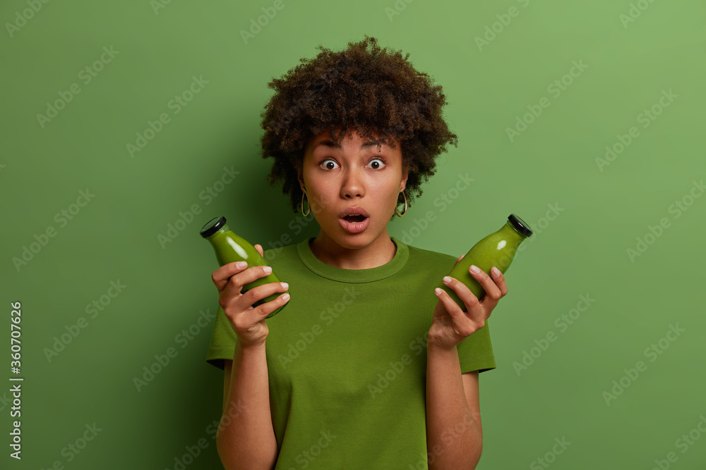 Attractive Afro American woman holds bottle of fresh blended green vegetable spinach smoothie, stares with shock, keeps to diet and has healthy nutrition, poses indoor. Vegetarian food concept - obrazy, fototapety, plakaty 