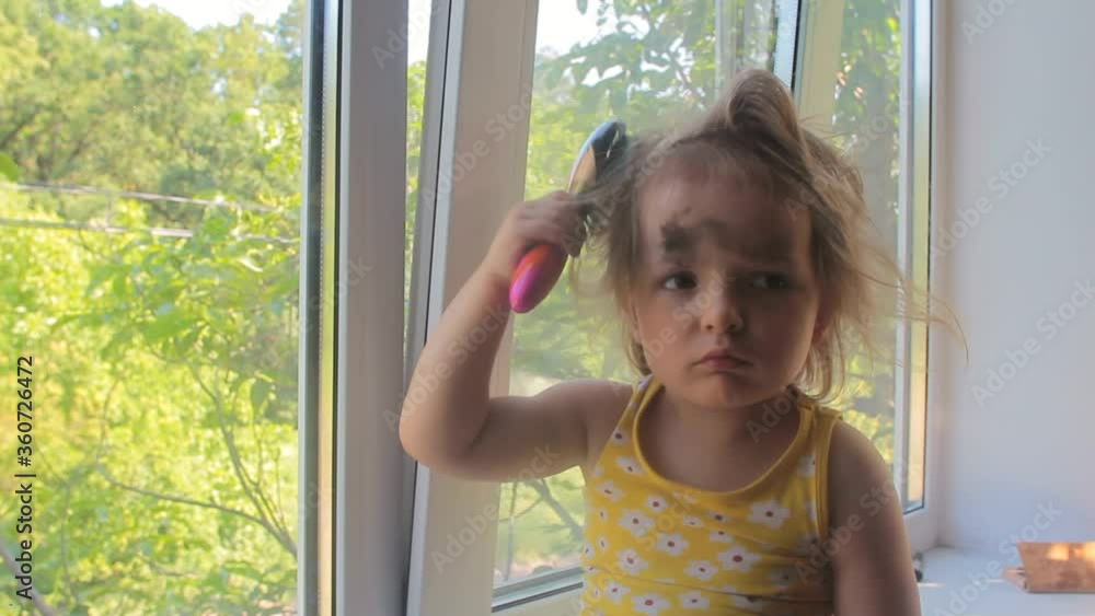 Child self care, little girl with eyeshadow on face brushing hair, sitting on window alone at home Child self care, little girl with eyeshadow on face brushing hair, sitting on window alone at home