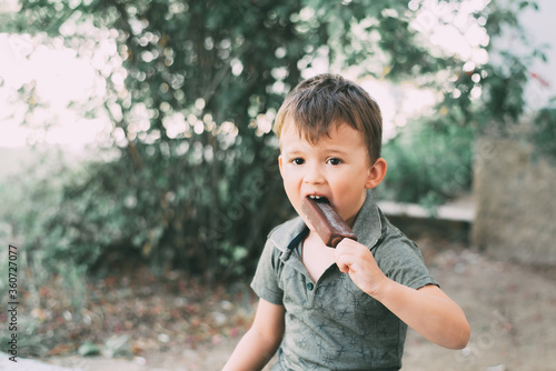 Boy eats ice cream on a stick covered with chocolate, outdoors © komokvm