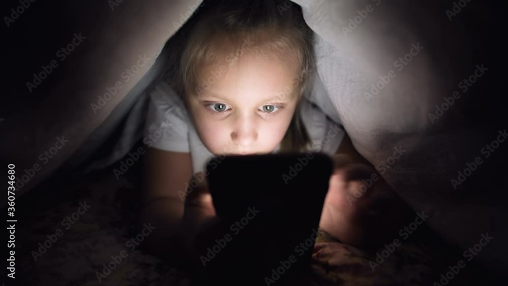 A keen little girl plays on a smartphone at night under a blanket. The child's departure from reality through mobile devices. Child uses secret phone A keen little girl plays on a smartphone at night under a blanket. The child's departure from reality through mobile devices. Child uses secret phone