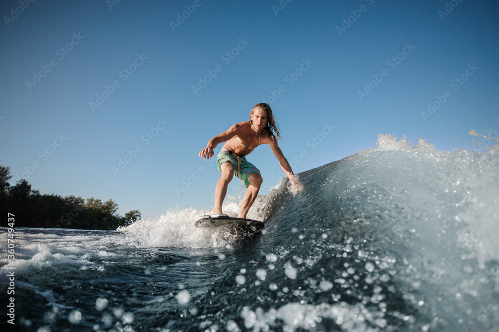 Tense and focused young man balancing on surfboard on river wave