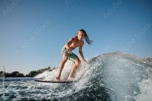 young active man rides on surfboard on wave against clear blue sky.