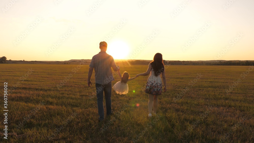 little daughter plays and jumps into field holding hands of mom and dad. healthy Happy family with child walks in park at sunset. baby and parent walk in sun. concept of happy family and childhood
