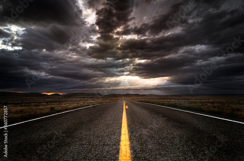 Lonely asphalt highway heading into a sunset with a cloudy sky and sun rays. There is a yellow painted lane in the middle of the road. No other cars can be seen. 