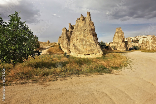 The amazing landscape of Cappadocia. A summer day, against the background of a cloudy sky and dried grass, yellow cliffs of a sandstone of a fanciful form rise. A dirt road winds between the rocks. 