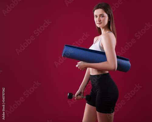 Sporty woman trainer in silhouette on a raspberry background. Photo of an attractive woman in fashionable sportswear. Girl with dumbbell and rug. Front view. Sport and healthy lifestyle