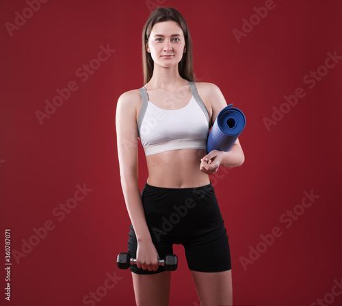 Sporty woman trainer in silhouette on a raspberry background. Photo of an attractive woman in fashionable sportswear. Girl with dumbbell and rug. Side view. Sport and healthy lifestyle