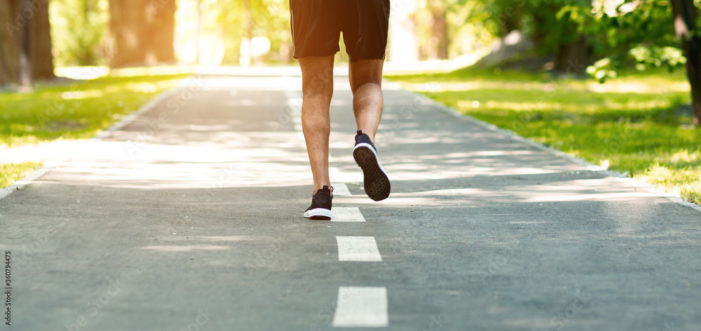 Unrecognizable millennial guy running along asphalt road at urban park, closeup of legs Unrecognizable millennial guy running along asphalt road at urban park, closeup of legs