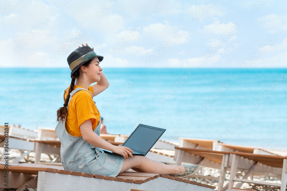 Freelance. A young woman sits relaxed on a sunbed with a laptop on her lap and looks out at the sea. Summer holiday. Copy space