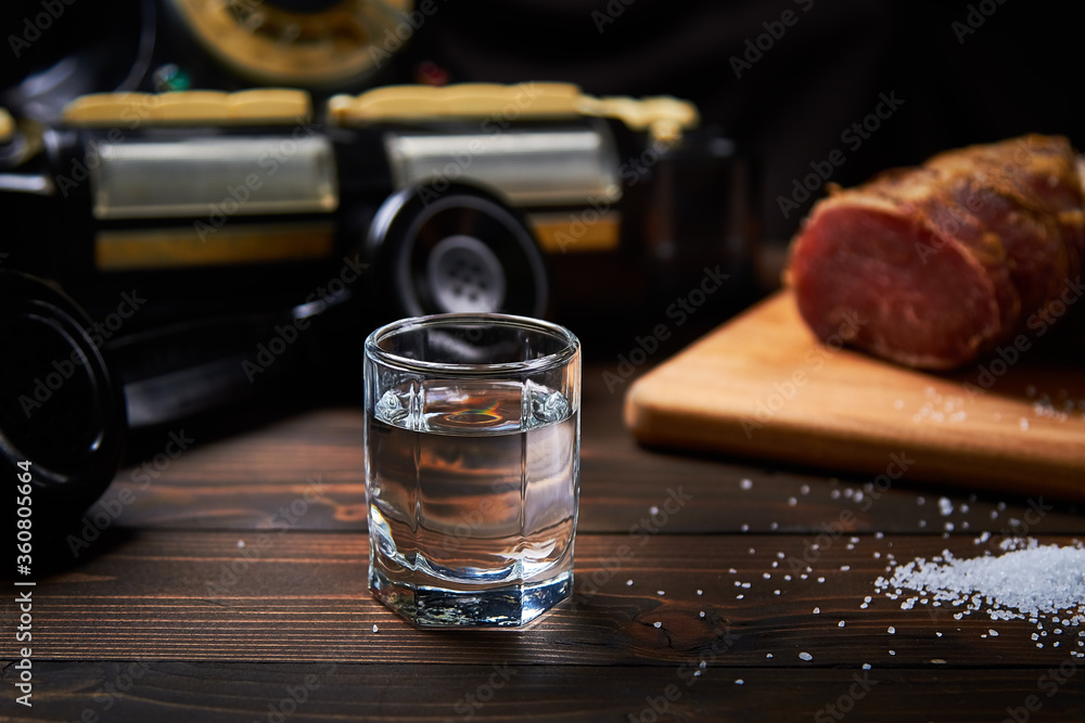 A vodka glass on an old table with an old black phone meat on the board with salt with shallow depth of field and blur.