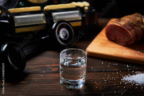 A vodka glass on an old table with an old black phone meat on the board with salt with shallow depth of field and blur.