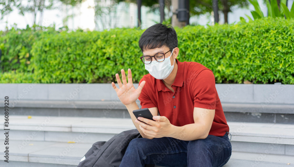 Fototapeta premium close up young asian man sitting at the park and wear face mask while using video call form smartphone to talking with friends and family for new normal and healthy lifestyle concept