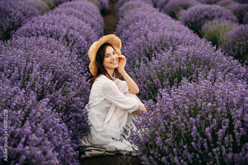 Young woman sitting between rows of lavender bushes, wearing a bohemian white dress and a straw hat, looking to camera.