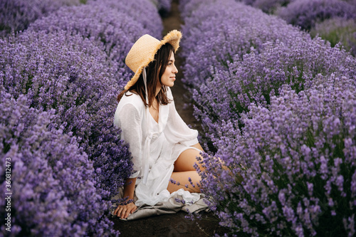 Young beautiful woman sitting in lavender field, wearing a bohemian white dress and a straw hat, looking to side.