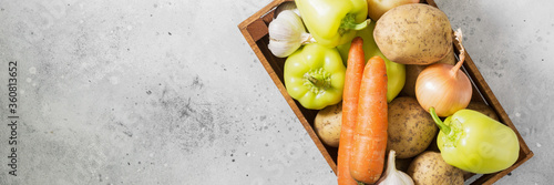 Various vegetables in a wooden box on a light gray table. Potatoes, peppers, onions, carrots and garlic in a brown wooden box. Top view with a space for text. Banner
