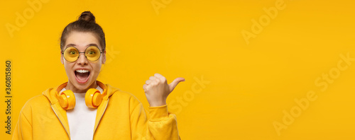 Horizontal banner of young excited girl wearing hoodie, glasses and wireless headphones around neck, pointing right to copy space, isolated on yellow background photo