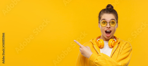 Horizontal banner of young shocked girl, wearing round glasses and headphones on neck, pointing left to copy space, isolated on yellow background photo