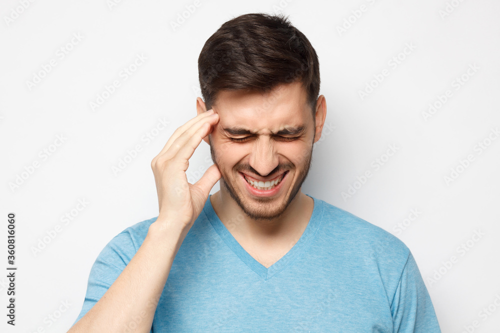 Headache concept. Young man touching temple with hand, trying to relieve pain, experiencing severe migraine, isolated on gray background Headache concept. Young man touching temple with hand, trying to relieve pain, experiencing severe migraine, isolated on gray background