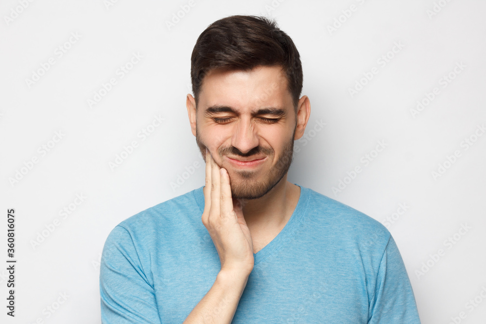 Young man with closed eyes suffering from severe toothache, touching jaw with fingers trying to ease strong tooth pain, isolated on gray background
