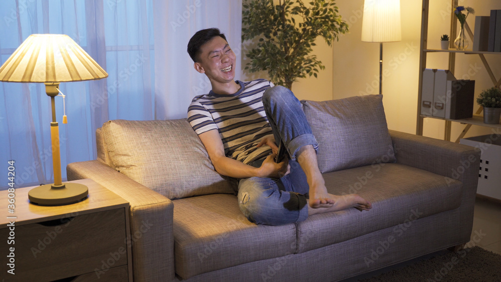 Obraz premium laid-back holiday asian guy changing tv channels found stand-up comedy and can't stop raising leg while laughing. korean male couch potato enjoying hilarious series at home alone before bedtime.