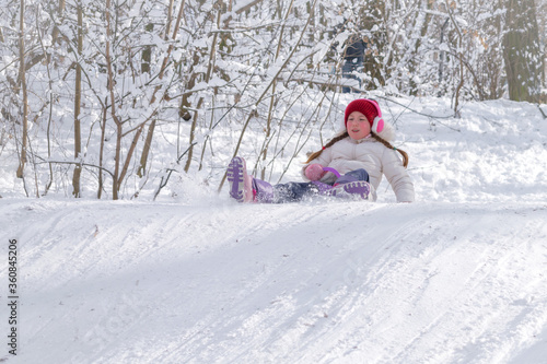 A child rides an iceboat in a snow-covered forest. A little girl sitting on her ass slides down a snow slide. Concept of children's winter active entertainment