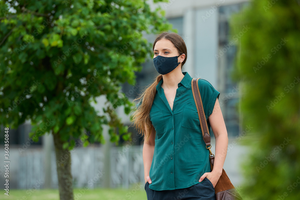Naklejka premium Woman in medical face mask walks between trees thrusting hand into pocket of trousers, holds a bag in downtown. Girl keeping social distance wears a protective mask to avoid the spread of coronavirus