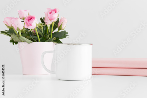 Enamel mug mockup with books and pink roses in a pot on a white table.