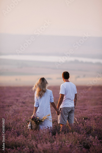 Loving couple admires the beautiful sunset in the lavender field. The guy and the girl are standing with their backs and holding hands.