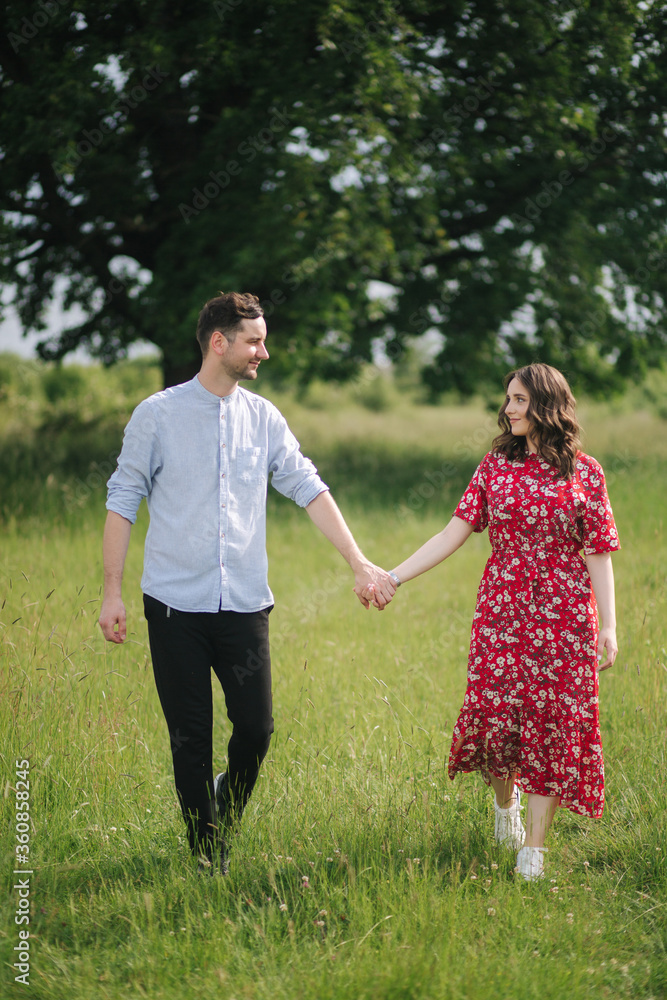 Young couple walking in front of big tree. Happpy man and woman spend time toghether. Woman in red dress Young couple walking in front of big tree. Happpy man and woman spend time toghether. Woman in red dress